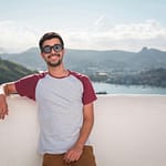 Young man in sunglasses leaning on a balcony with a scenic city view in the background.